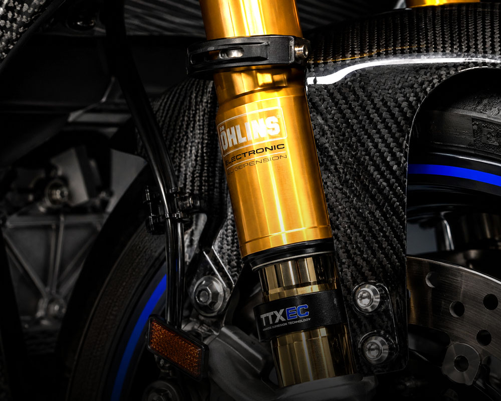 Macro photograph of Öhlins gold-colored electronic suspension fork on a superbike, carbon fiber front fender visible in background, studio lighting, crisp detail, product photography style